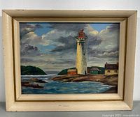 Framed lighthouse painting showing a rocky shore with lighthouse and adjacent buildings under a partly cloudy sky, visible minor wear on the frame.