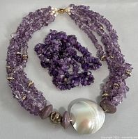 Four strands of polished amethyst chips with metallic and pearl-like accents and large round white shell centerpiece, laid flat on gray background.