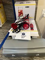 Bella immersion blender in red, power cord visible, placed in front of its box and instruction manual with a gray plastic case nearby