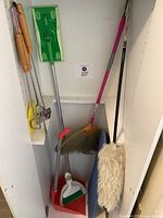 Shelf with various cleaning items including a red dustpan with green brush, broom with pink handle, mop duster with white head, broom with yellow bristles, and long handle fly swatters.