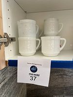 Seven white milk glass mugs with floral embossed pattern shown stacked inside a cabinet.