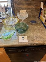 Photo of all six glass items arranged on countertop: includes clear celestial footed bowl, clear hobnail cake plate, green mixing bowl, clear scalloped bowl with blue center, green wavy-edged bowl, and clear pressed glass plate.