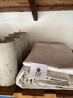 Photo showing four cylindrical concrete counterweights with metal eyelets and two folded white canvas tarps on wooden shelves.