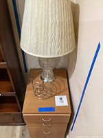 Photo showing the crystal lamp with fabric lampshade, wire waste basket, and three drawer rolling filing cabinet with metal handles from front angle.