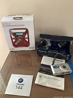 Etan FRX3 weather radio in box alongside Panasonic Lumix FX3 digital camera with cables, instruction manual, and boxes on glass table.