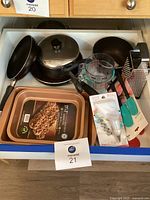 Drawer with various kitchen items including two copper nonstick baking pans, two soup pots with lids, two Pyrex measuring cups, a metal cheese grater, a pastry bag with piping tips, and two teal-handled spatula sets.