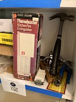 Photo showing Flamebuster fire extinguisher box, black handled hammer, and some hardware items including a white electrical switch and blue pliers.