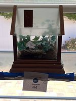 Glass and wood terrarium with faux greenery inside, shaped like a small house with pitched roof and brown wood trim