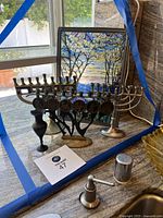 Three brass menorahs and a decorative window panel placed together on countertop with natural light from window behind.