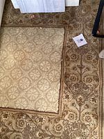 Partial view of rug showing beige central area with geometric floral pattern and brown ornate border with floral motifs; dirt and wear visible; edges slightly worn.