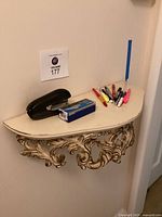 Mounted cream-colored half-moon wall table with ornate carved gold-tone molding underneath, some damage visible. Black stapler, box of staples, and assorted pens on top.
