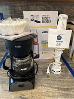 Mr. Coffee coffee maker with glass carafe, cord coiled around base, Bella Rocket blender box, and a white Braun hand mixer.