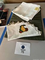 Four dark green placemats stacked with five cream-colored embroidered Lenox napkins with pear and berry design and scalloped edges on a table surface.