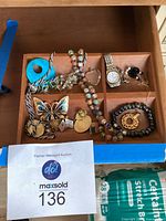 Top view of assorted costume jewelry in a wooden box divided into compartments showing beaded bracelets, charms, a butterfly brooch, and the watch.