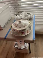 Octagonal vegetable tureen with gold handles and floral design placed on table with cloth, along with round serving tray and other china pieces, showing overall visual condition and arrangement.