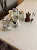 Overview of all salt, pepper, sugar, and honey dispensers on a white countertop, showing variety of materials including wood, glass, crystal, and plastic.