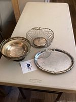 Three items on a white table: silver plated oval serving plate, metal apple-shaped dish, and silver plated bowl with handle.