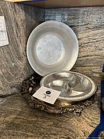 Large silver plated serving dish with three ornate flower feet, plain silver plate behind it, all showing wear and patina.