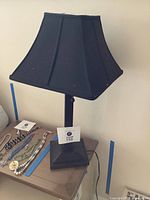 Full view of black metal table lamp with square black fabric shade on a wood surface; lamp has square base and no bolt on shade.