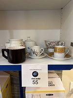 Shelf with seven teacups and saucers of various designs including one music box teacup and one brown mug without saucer