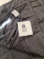 Close-up of blanket fabric, showing dark gray quilted squares and two tags: one brand tag 'marari eden' and one lot tag 'masold 107'.