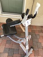 Ab Coaster exercise machine angled view showing padded seat, seat supports, handlebars with foam grips, and plastic protective wrappings on key parts.