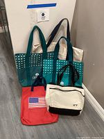 Four tote bags displayed against a wall: two green bags with circular logos, one red bag with American flag, one white and black bag with ARY logo
