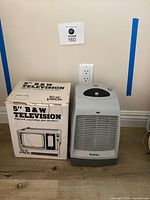 Vintage 5 inch black and white Tote Vision television in original box next to a Holmes floor heater placed against a wall on wood flooring.