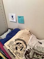 Stack of 10 assorted women's vintage T-shirts in various colors and graphic prints, showing wear and wrinkles, folded on a flat surface against a white wall.