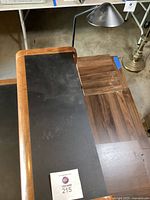 Wooden desk with black matte surface panel and low wooden shelf cabinet beside it. Black reading lamp placed on thin metal table behind desk.