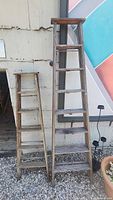 Photo showing both antique wooden stepladders side by side against a wall, longest measures about 8 feet.
