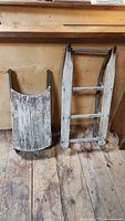 Two wooden sleighs placed upright against a wooden wall and floor, one ladderr style and one flat riding surface style.