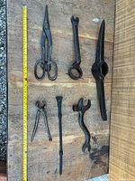 Overview of eight antique metal tools of various types arranged on wood. Includes scissors-style tin snips, multi-wrench, hook, and tongs. Shows size, shape, and signs of use.