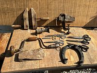 Photo of assorted vintage tools including vices, hand drill, scissors, cleaver and horseshoe on a wooden surface in daylight.