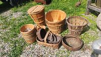 Photo shows a group of seven baskets of various types in natural outdoor lighting on gravel, including wooden bushel basket, tall lidded wooden basket, woven basket, and handled wooden baskets.