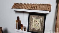 Wooden cigar mold leaning against wall, with three vintage bottles and framed embroidery below it.
