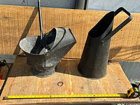 Photo showing two metal coal scuttles and a metal pail arranged on a wooden surface with measuring tape for scale. The items are black and show wear consistent with age.