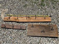 Three wooden boards of varying sizes laid out on gravel; each has multiple black metal double-prong coat hooks mounted.