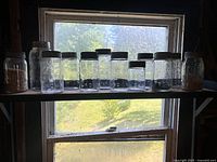 View of a shelf holding ten vintage Crown glass sealer jars of various sizes with metal lids, some containing remnants of dried food material.