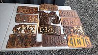 Top-down photo showing a collection of 13 old Ontario license plates on a table, some heavily rusted, including a yellow and black 1962 plate C 325.