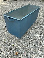 Side view of a rectangular metal ice tub painted dark blue with a side handle, placed on gravel ground outdoors.