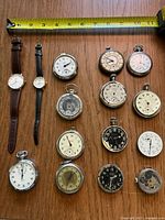 Full view of collection of 14 watches including 2 wristwatches, pocket watches of various dial conditions, and a stopwatch, arranged under measuring tape showing size approx 4 inches.