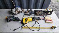 Lot of power tools on white table showing circular saw, router, bench grinder, reciprocating saw with cord, tape measure, and small hardware box.