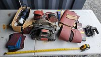 Wide view of the main lot items showing electric belt sander, orbital sander, sanding belts, sanding pads, electrical cords, and measuring tape for scale.