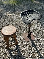 Two stools side by side on gravel surface: a small wooden stool and a larger industrial style metal stool with tractor seat.