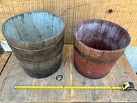 Two wooden water pails side by side on a wooden table with measuring tape laid out in front for scale. Both have metal bands; one is natural wood color, the other red painted.