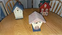 Three hand-painted birdhouses displayed on a wooden table showing front and side views.