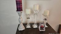 Four decorative candle holders displayed on a dark surface against a white wall, including one cream-colored ornate holder and three clear glass faceted holders, some holding candles.