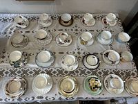 Overview photo of 19 cups and saucers arranged on patterned tablecloth showing variety of floral and gold trimmed sets.