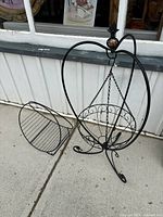 Two black metal items on a concrete floor by a building wall: a spherical hanging plant stand suspended by chains from a curved frame with scrollwork, and a detached half-circle magazine holder with wire rack design.